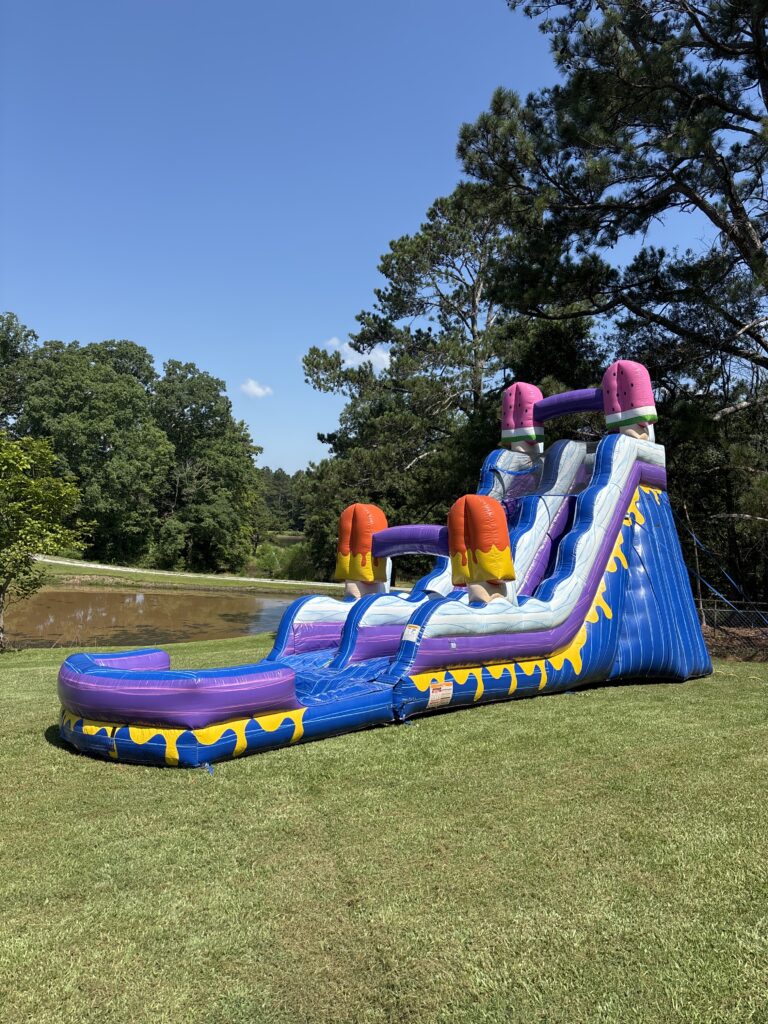 Colorful inflatable water slide set up on a grassy lawn near a pond, surrounded by tall trees and a clear blue sky. The slide features vibrant purple, blue, and yellow colors with two large ice pop designs at the top and two sliding lanes curving into a splash pool at the bottom.